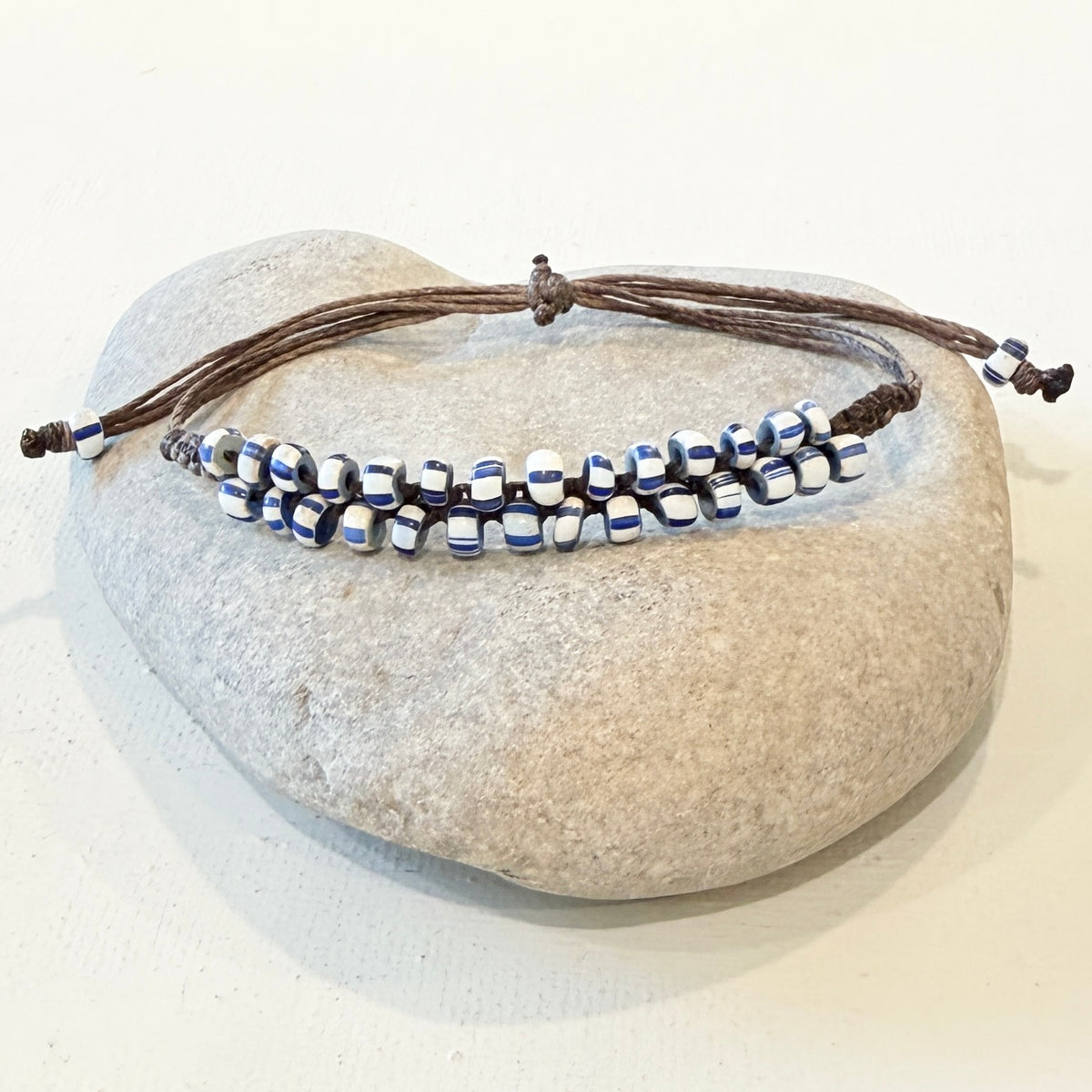 A beaded adjustable cord bracelet with blue and white striped beads and dark brown adjustable cords, displayed on a smooth light gray stone against a white background.