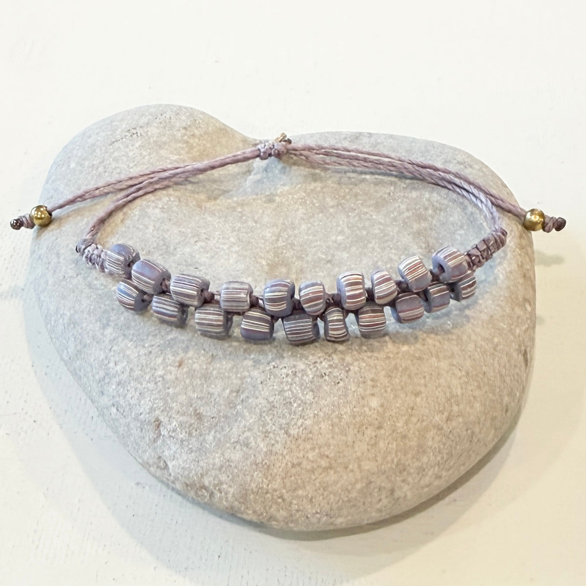 A beaded bracelet with striped lavender, purple, and gray beads on a braided cord, displayed on a smooth light gray stone against a white background.