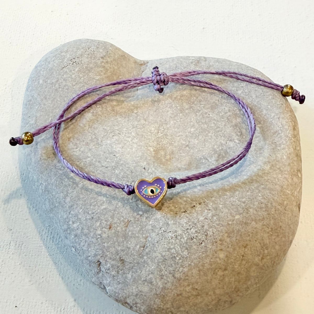 A purple braided cord bracelet with a small heart-shaped evil eye charm, displayed on a smooth, light gray stone against a white background.