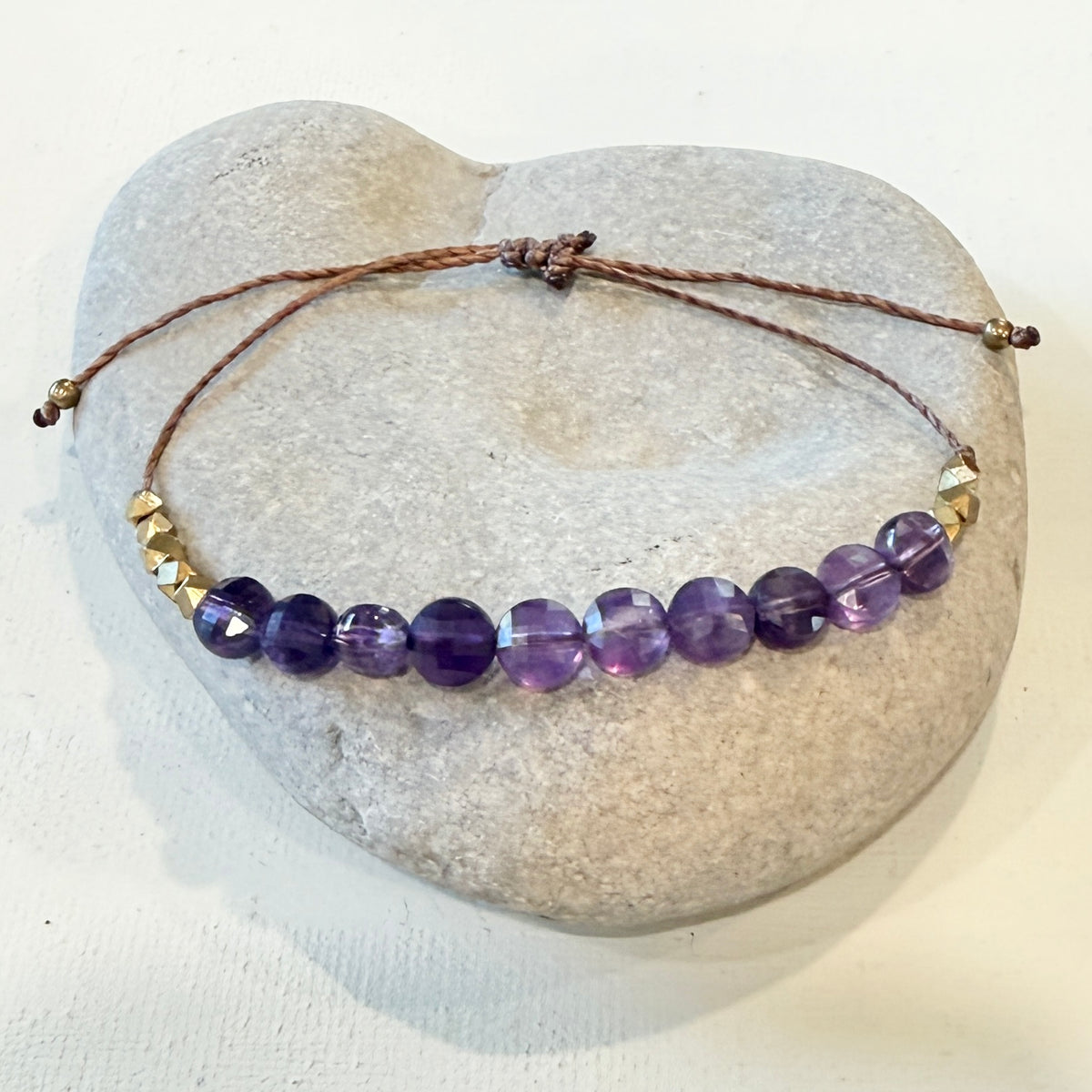 A beaded adjustable cord bracelet with faceted purple amethyst beads and small gold spacer beads, displayed on a smooth light gray stone against a white background.