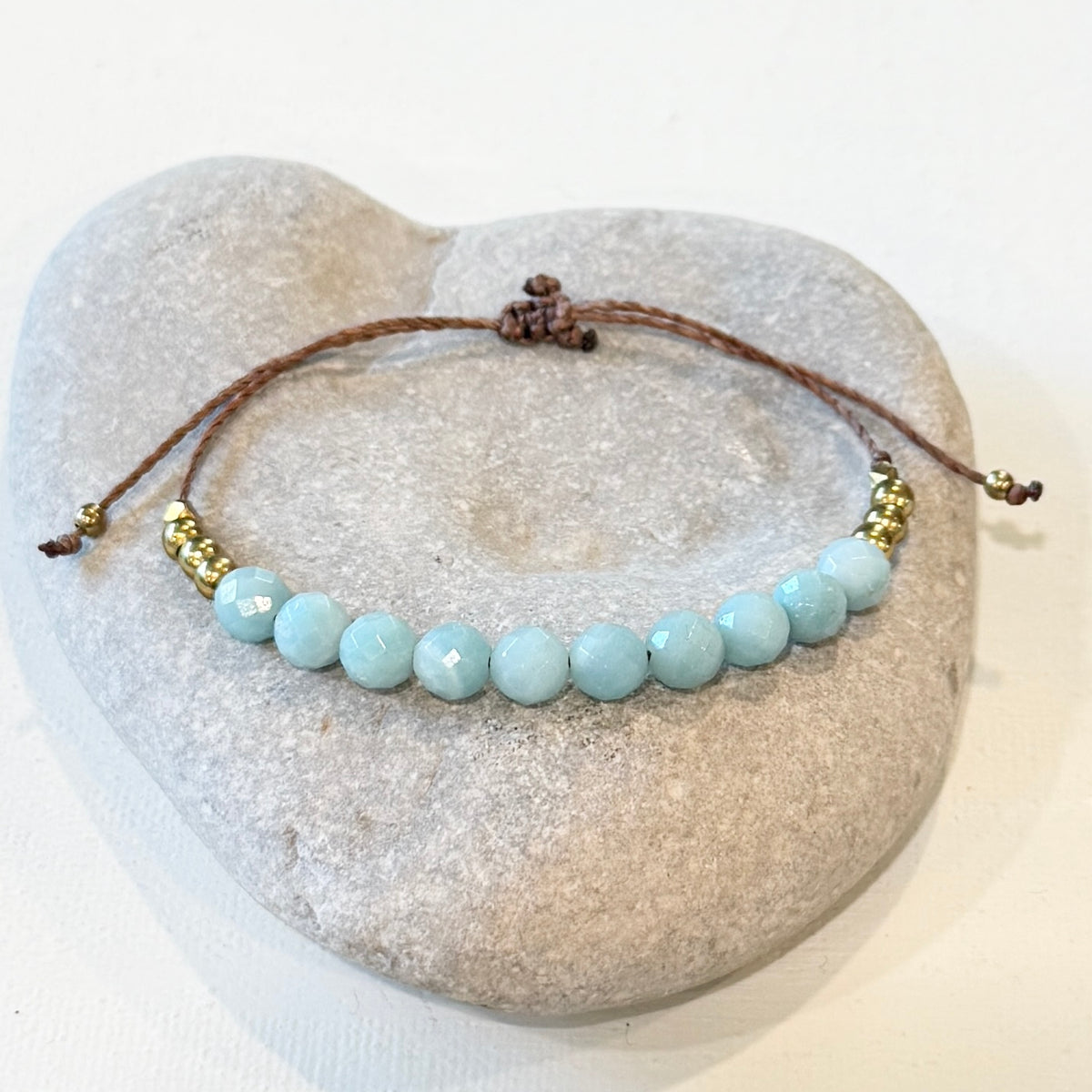 A beaded cord bracelet with light blue faceted beads and small gold spacer beads, displayed on a heart-shaped stone stand against a white background.