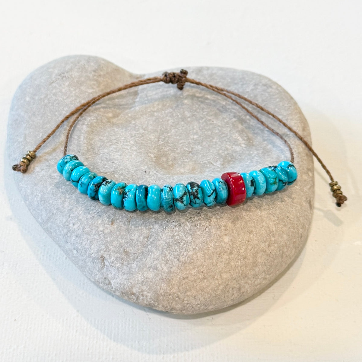 A beaded adjustable cord bracelet with turquoise-blue round beads and one red accent bead, displayed on a light gray stone prop against a white background.