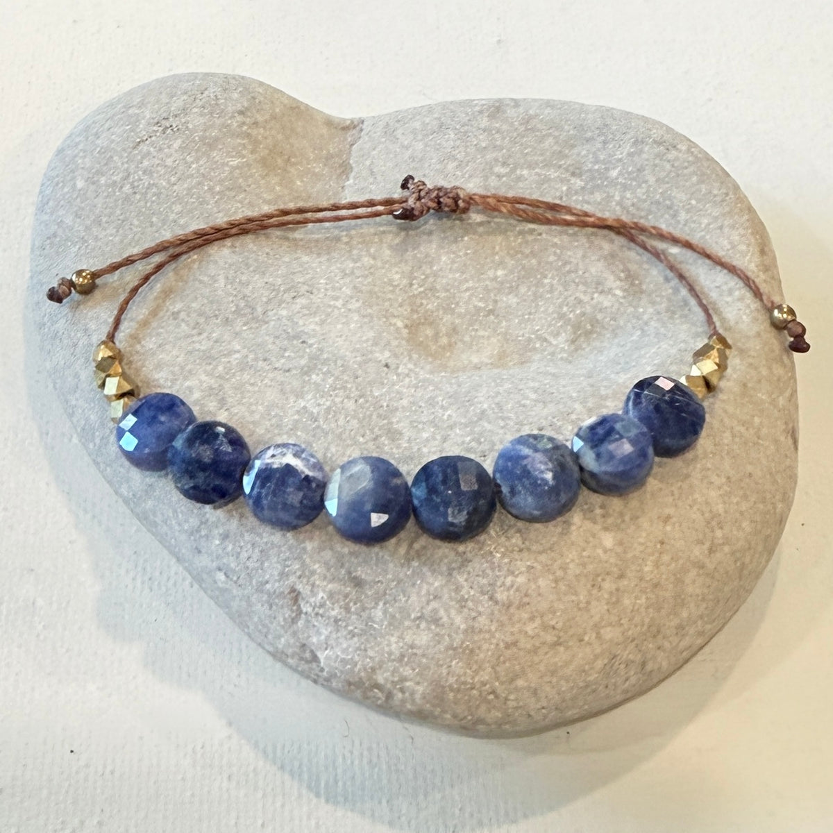 A beaded bracelet with faceted dark blue sapphire-like beads and small gold spacer beads on a brown cord, displayed on a smooth light gray stone against a white background.