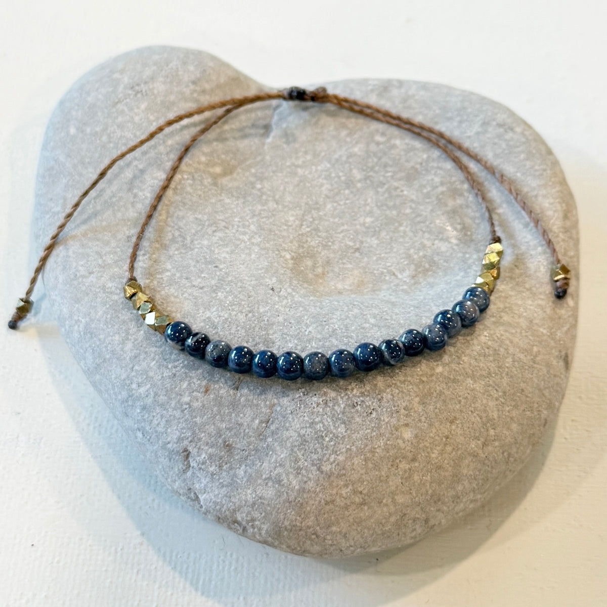 A beaded necklace with dark blue round beads and small gold-toned spacer beads on a brown cord, displayed on a smooth gray stone against a white background.