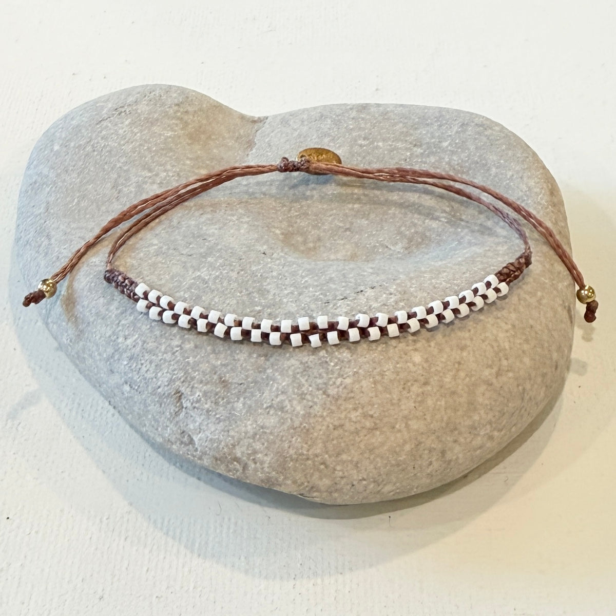 A beaded adjustable cord bracelet with white and dark brown beads, displayed on a light gray stone stand against a white background.