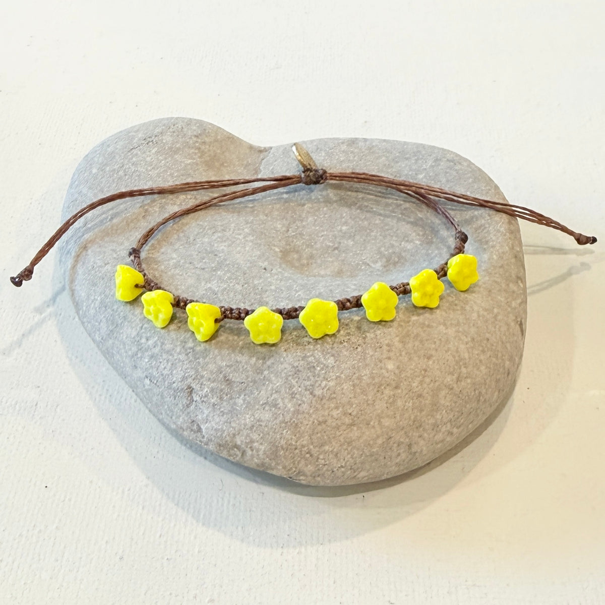 A brown cord bracelet with seven small yellow flower beads, displayed on a smooth gray stone against a white background.