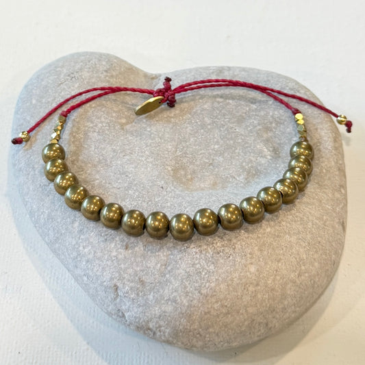 A beaded bracelet with metallic bronze beads and a red adjustable cord closure, displayed on a light gray stone surface.