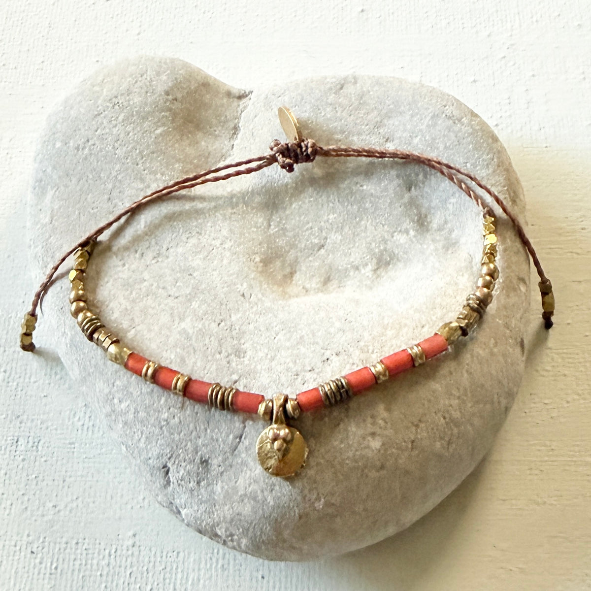 a light color stone with a bracelet displayed on top with cord, red and gold beads with a gold color talisman on an off white background.