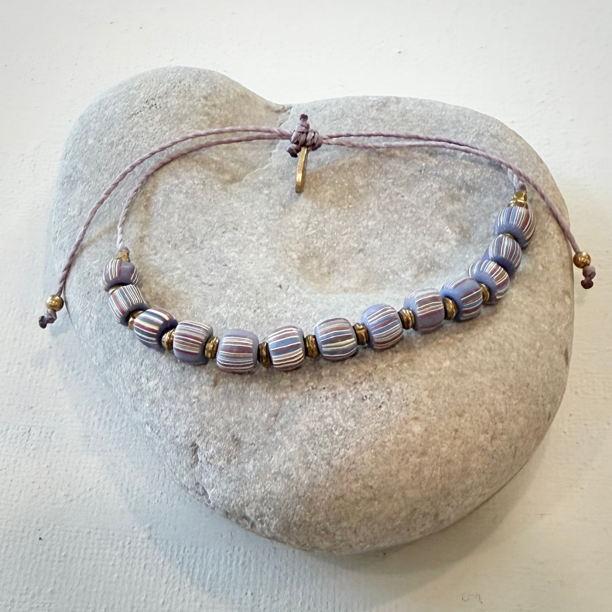 A beaded necklace with striped lavender, purple, and white glass beads, displayed on a light gray stone stand against a white background.