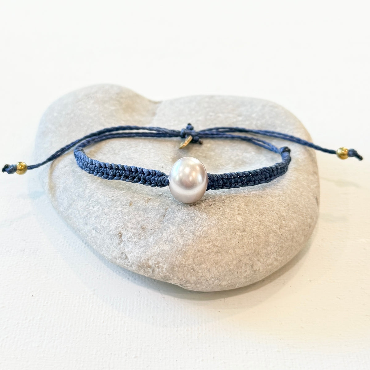 a bracelet on navy cord  with a pearl and brass beads sitting on a grey rock with a white background. 