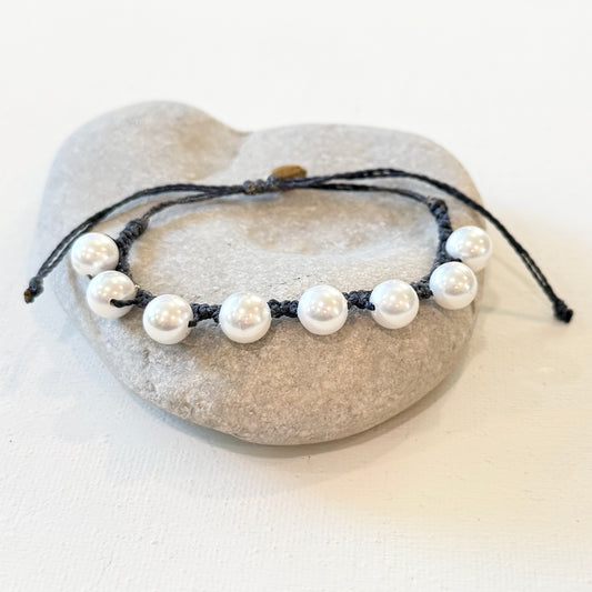 A beaded bracelet with white pearl-like beads on a black cord, displayed on a light gray stone stand against a white background.