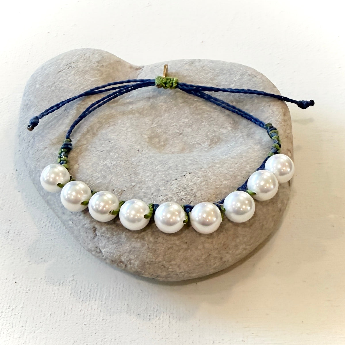 A beaded necklace with large white pearls strung on a navy blue cord with small green accents, displayed on a smooth gray stone against a light background.