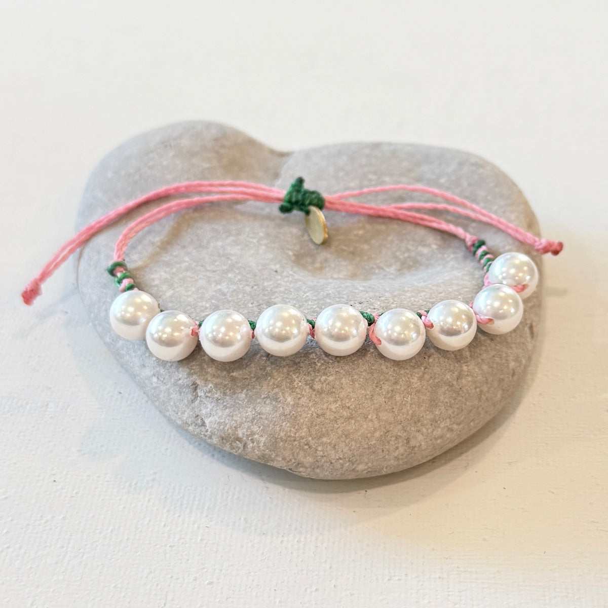 A beaded bracelet with white pearl-like beads threaded on a pink cord with small green accents, displayed on a smooth gray stone against a light neutral background.