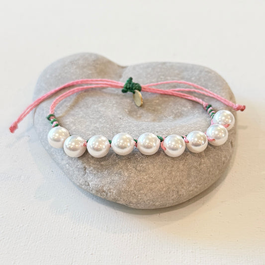 A beaded bracelet with white pearl-like beads threaded on a pink cord with small green accents, displayed on a smooth gray stone against a light neutral background.