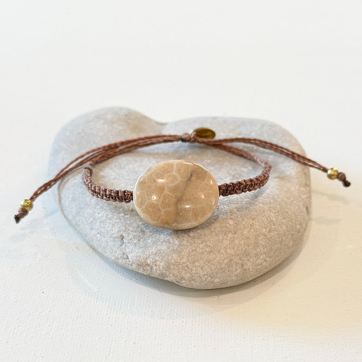 A brown cord bracelet with a round beige stone centerpiece, displayed on a smooth light gray stone against a clean white background.