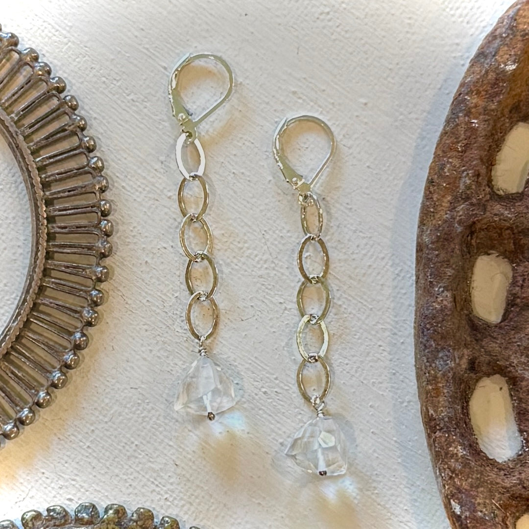 A pair of dangling earrings with textured silver-tone chain links and clear faceted crystal drops, displayed on a textured white surface with rustic decorative props.