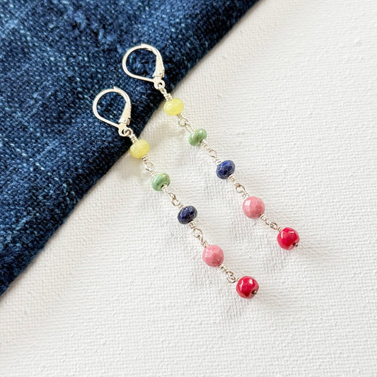 Multicolored beaded earrings on a denim and white fabric background. Gemstones ranging from coral, rhodochrosite, lapis lazuli, turquoise and apple jade.