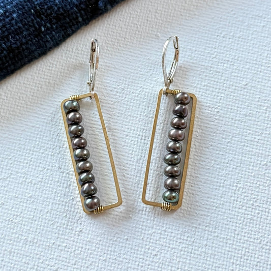 Pair of earrings with rectangular gold frames and dark fresh water pearl  beads on a light fabric background with sterling silver ear wires.