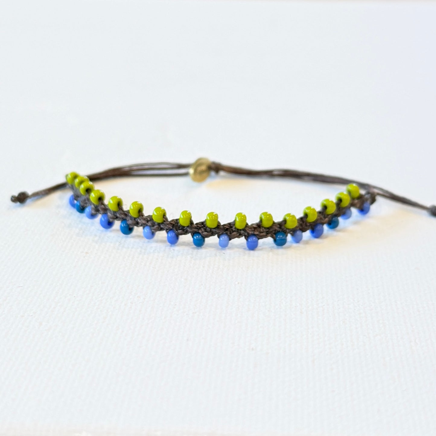 blue and green tiny seed beads linked with chocolate brown waxed nylon. Adjustable in size with a brass monogram tag.
