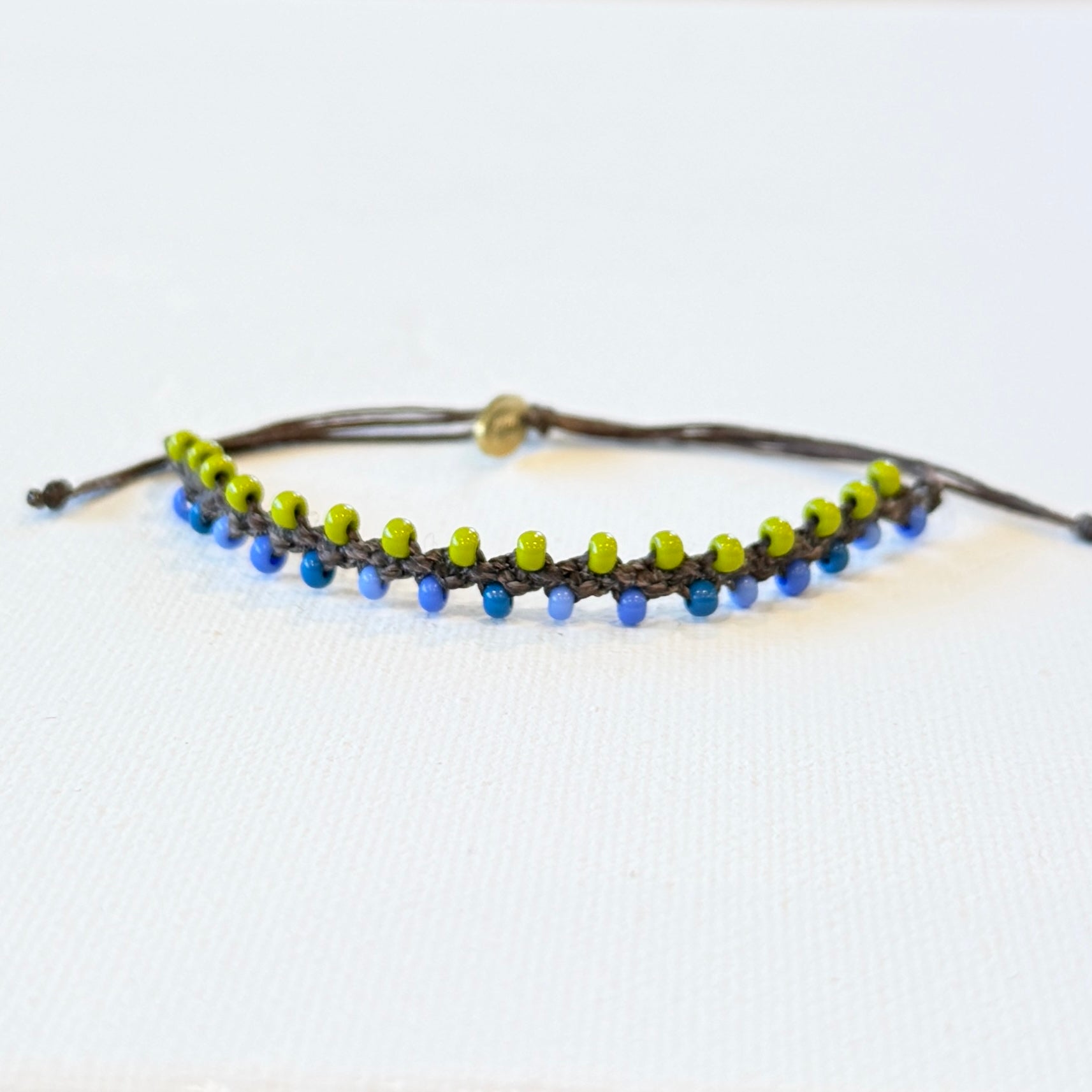 blue and green tiny seed beads linked with chocolate brown waxed nylon. Adjustable in size with a brass monogram tag.