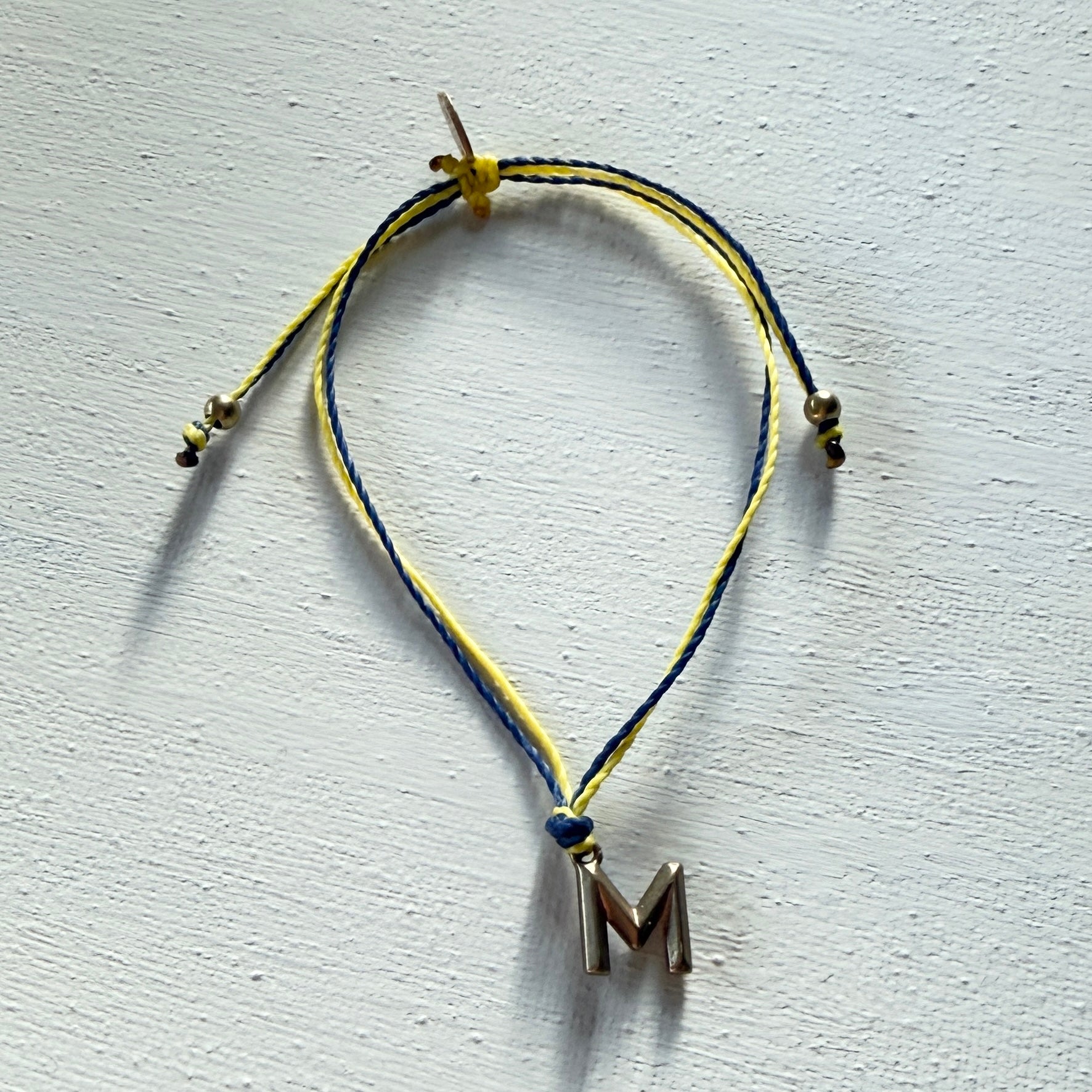 Bracelet with yellow and blue cord and letter 'M' charm on a light gray background