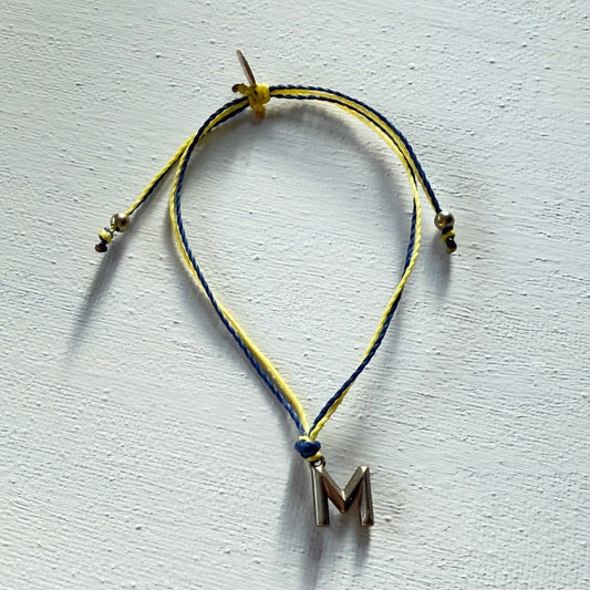 Bracelet with yellow and blue cord and letter 'M' charm on a light gray background