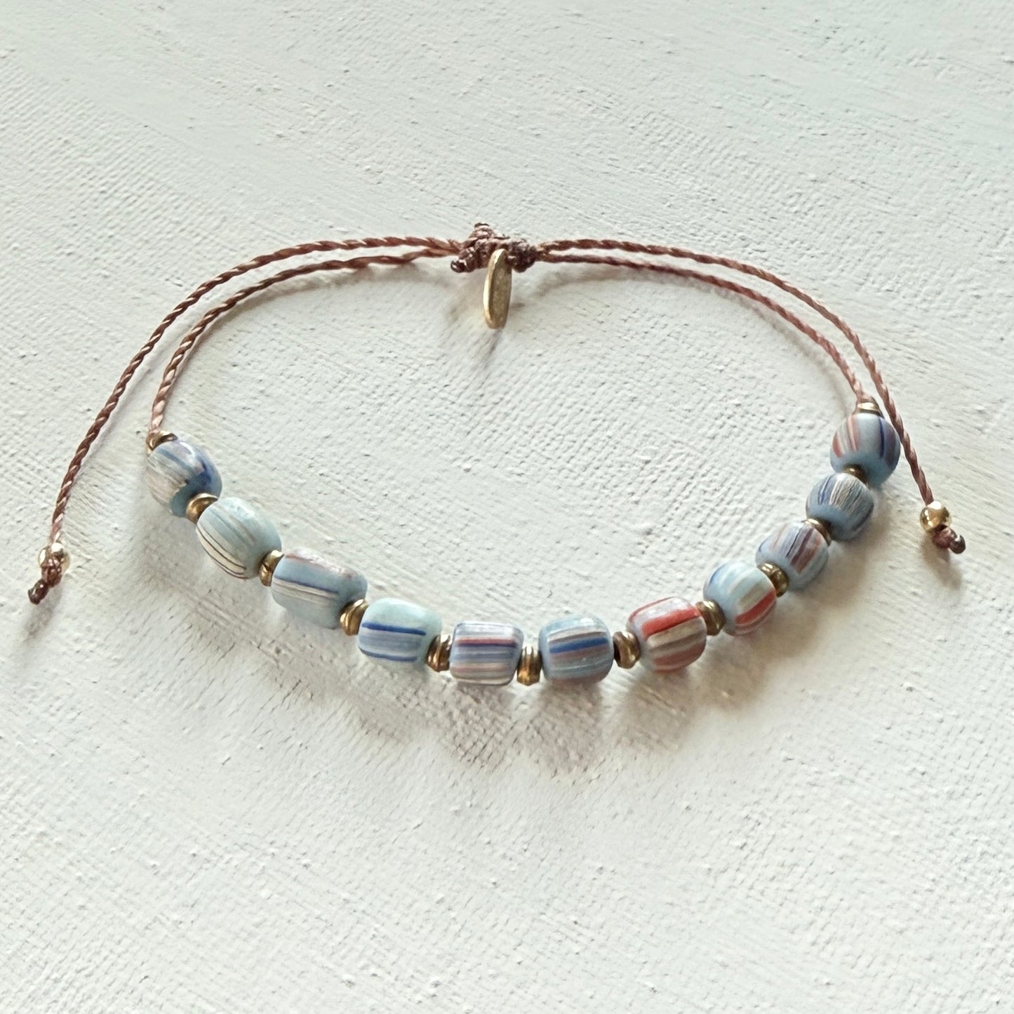 Beaded bracelet with blue striped beads on a brown cord with brass beads on a textured white surface