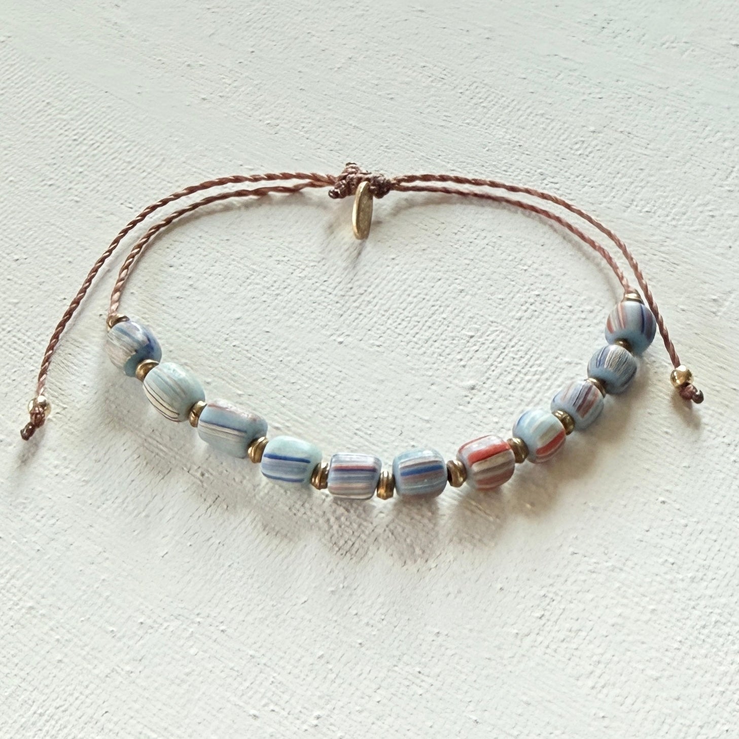 Beaded bracelet with blue striped beads on a brown cord with brass beads on a textured white surface