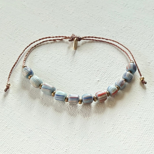 Beaded bracelet with blue striped beads on a brown cord with brass beads on a textured white surface